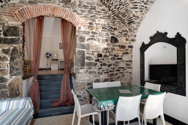 A stone-walled room with a dining table, white chairs, a TV, and a staircase leading to another room.
