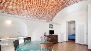 Un coin repas avec une table recouverte d’une nappe verte, des chaises en bois, un plafond en briques et une cuisine en arrière-plan.