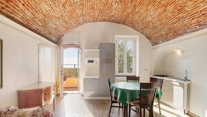 Une pièce avec un plafond en briques, une table à manger avec des chaises, un canapé, un coin cuisine et une vue sur l’extérieur, visible à travers une porte en arc de cercle.