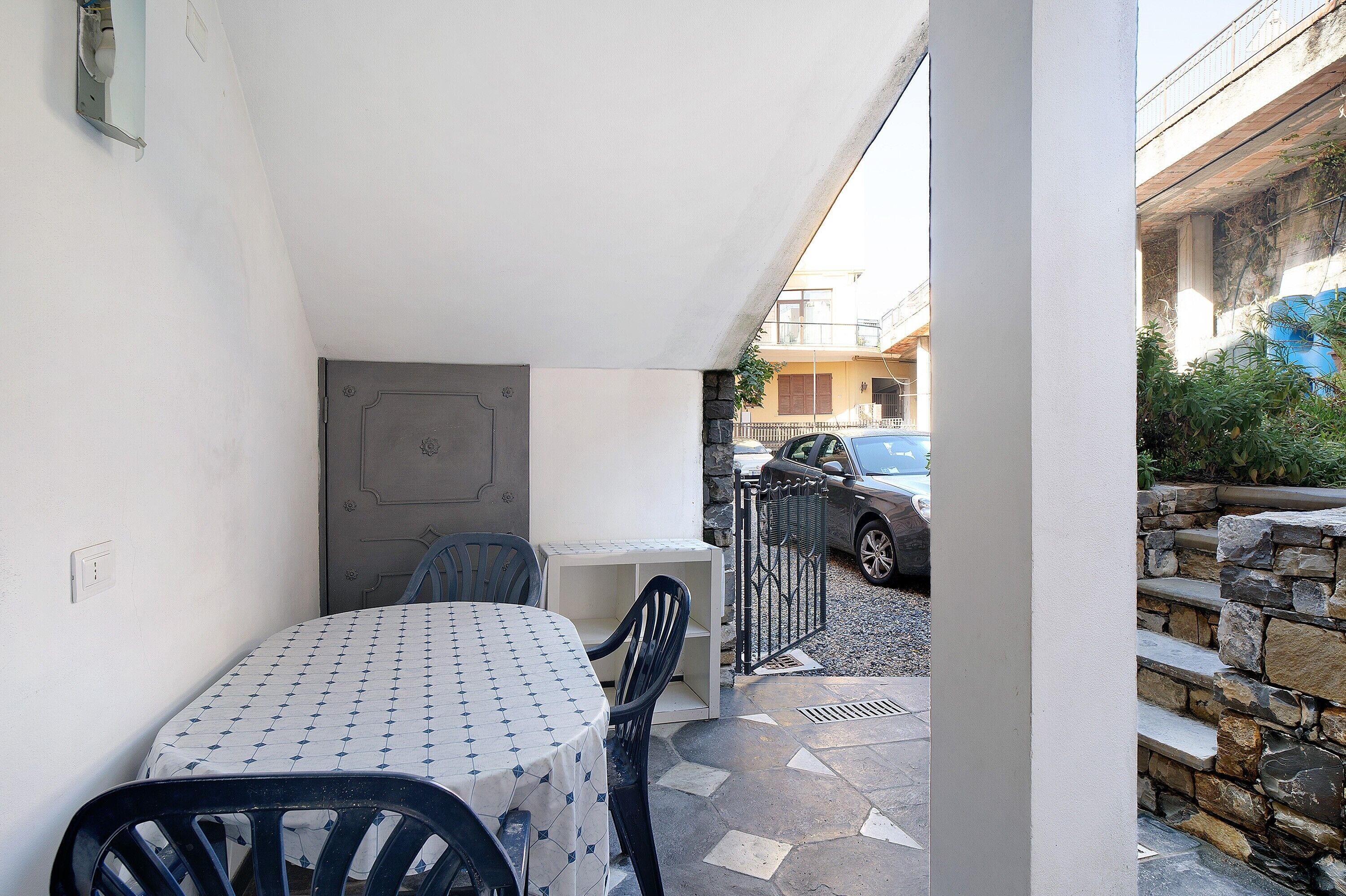 Un pequeño espacio al aire libre con mesa y sillas, una puerta y vista a una calle con un coche estacionado.
