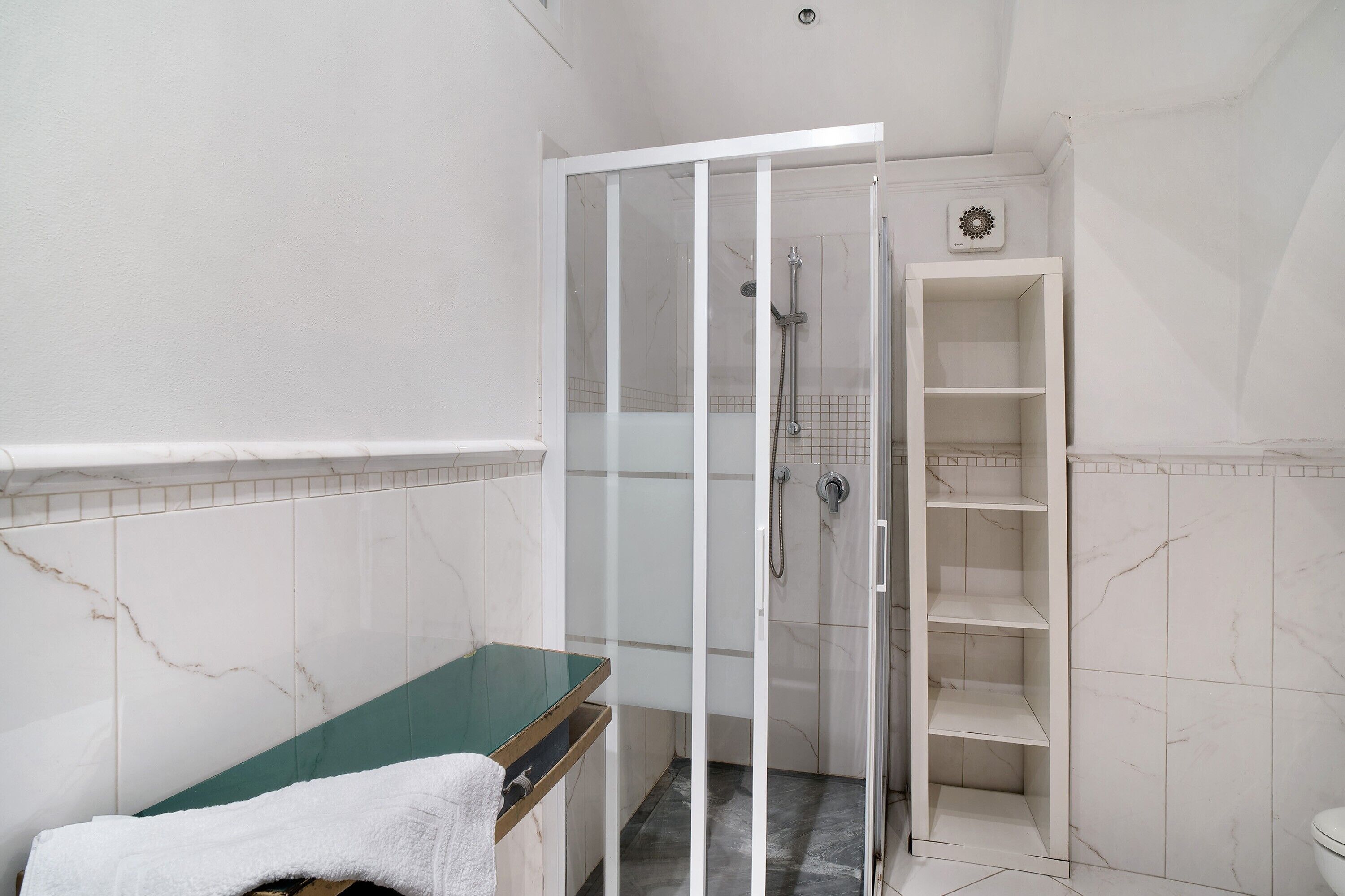 Ein Badezimmer mit einer einachigen Duschkabine, einer Wand mit Marmorplatten und einer weißen Ablage.