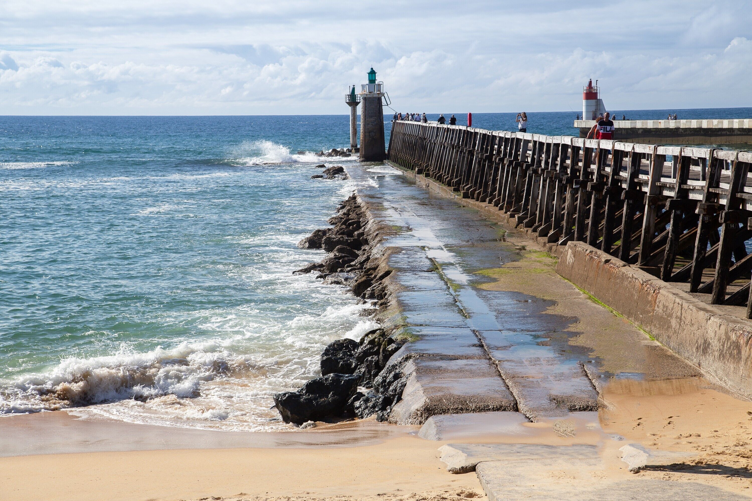 Ein Küstensteg mit einem Leuchtturm, Wellen schlagen an den Felsen, und ein Sandstrand.