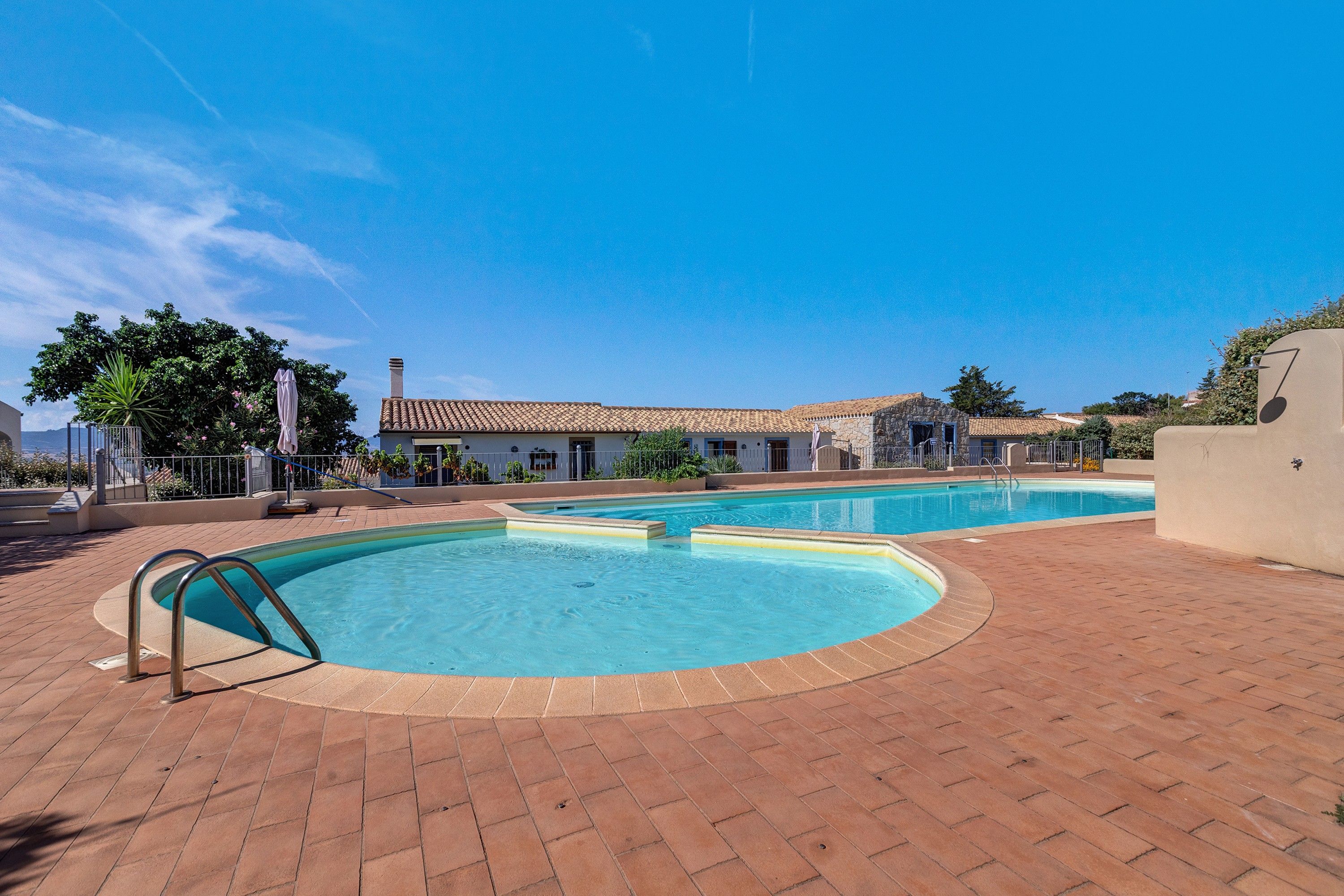 A swimming pool area with two pools, a covered patio, and a building with a tiled roof.