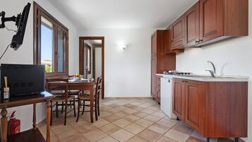 A compact kitchen with wooden cabinets, a dining table with chairs, a small refrigerator, and a window with a view.