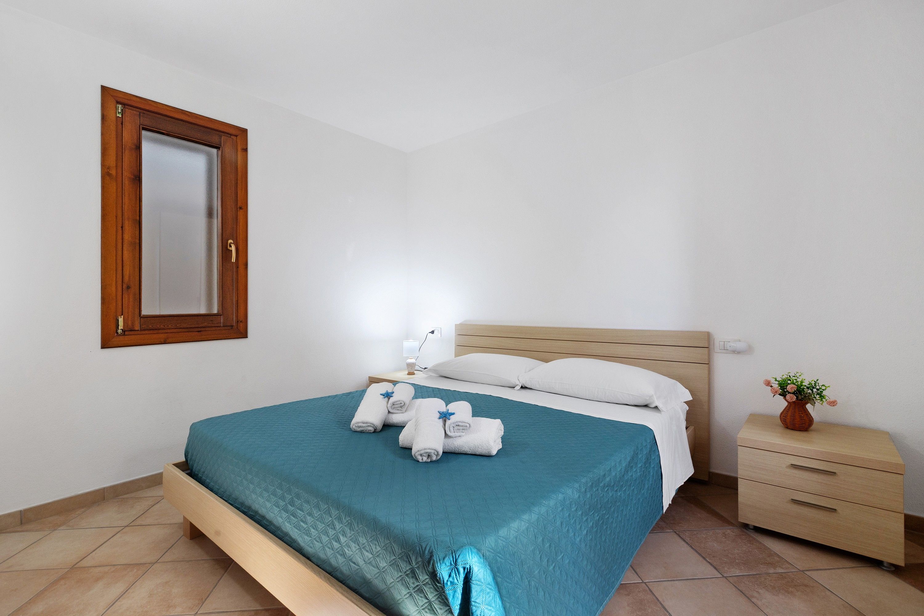 Een netjes opgemaakt bed met een blauwgroene spreidek en witte kussens, een houten nachtkastje met een bloevas en een klein dressoir aan de rechterkant.