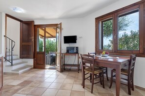 Un coin repas avec une table et des chaises en bois, un téléviseur posé sur un meuble, et une fenêtre donnant sur un paysage verdoyant.