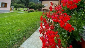 Un edificio con balcón de madera, un camino de piedra y un patio césped con flores rojas.