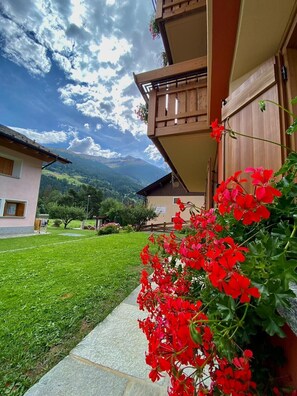 A building with a wooden balcony, a stone pathway, and a grassy yard with red flowers. - Holiday Apartment "Casa Clus Bormio 1" with Mountain View, Private Garden & Wi-Fi (Valdisotto)
