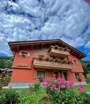 Un bâtiment sur lequel on peut lire l’inscription « Casa Clus », des balcons ornés de jardinières fleuries et un jardin où poussent des fleurs roses.