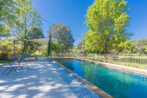 Una piscina rectangular con muelle de madera y piedras blancas, rodeada de árboles y una valla.