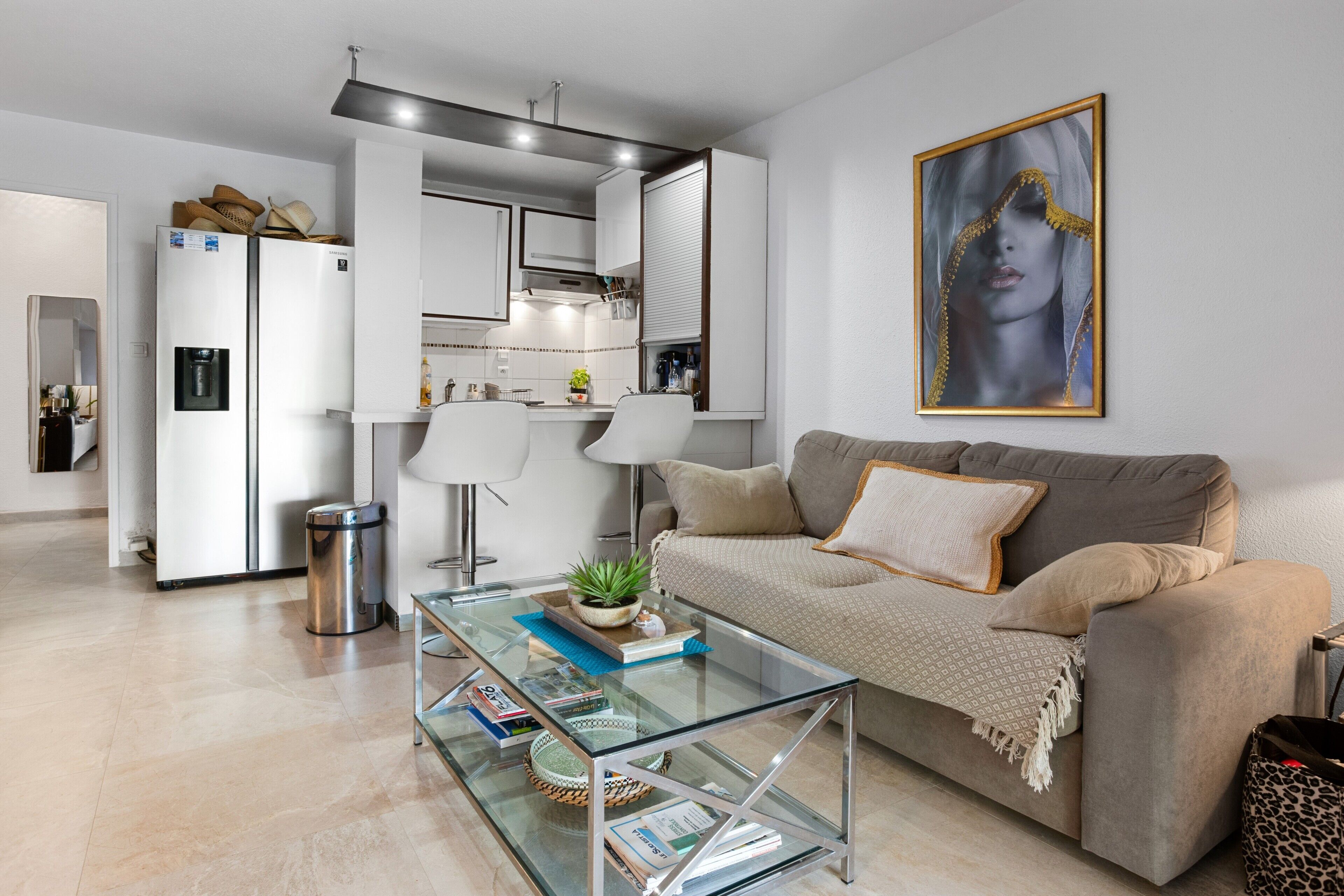 A modern living room with a glass coffee table, beige sofa, and a kitchen area with white cabinets and a refrigerator.