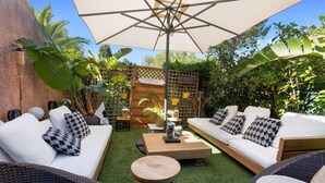 Une terrasse extérieure avec deux canapés blancs, une table ronde en bois et un grand parasol qui offre de l’ombre.