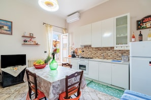 Uma cozinha com armários brancos, painel de azulejos, uma mesa de jantar com toalha de mesa estampada e uma TV sobre um suporte.