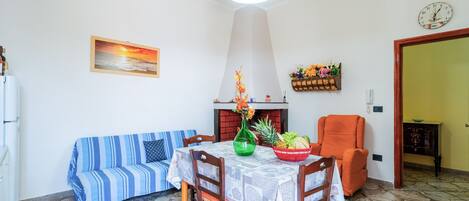 A dining area with a table set for two, a sofa, a chair, and a fireplace.