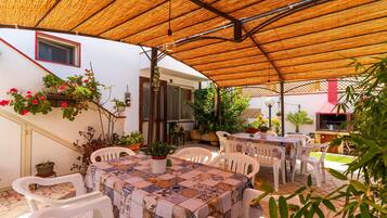 Eine Terrasse im Freien mit einem Tisch, bedeckt mit einer gemusterten Tischdecke, umgeben von weißen Plastikstühlen.