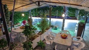 Area pranzo all'aperto con sedie e tavoli in plastica bianca, circondata dal verde e vista su un corso d'acqua.