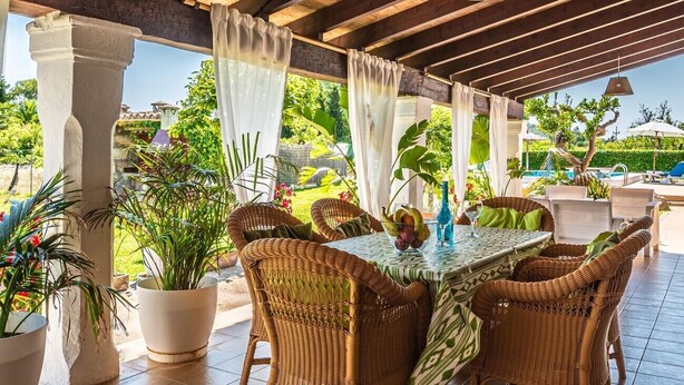 Une terrasse extérieure avec des meubles en osier, une table sur laquelle est posé un panier de fruits et des plantes en pot.