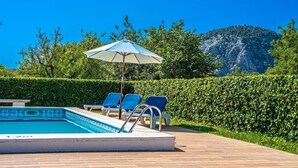 Une piscine avec des chaises longues et un parasol, entourée d’une haie et avec une montagne en arrière-plan.
