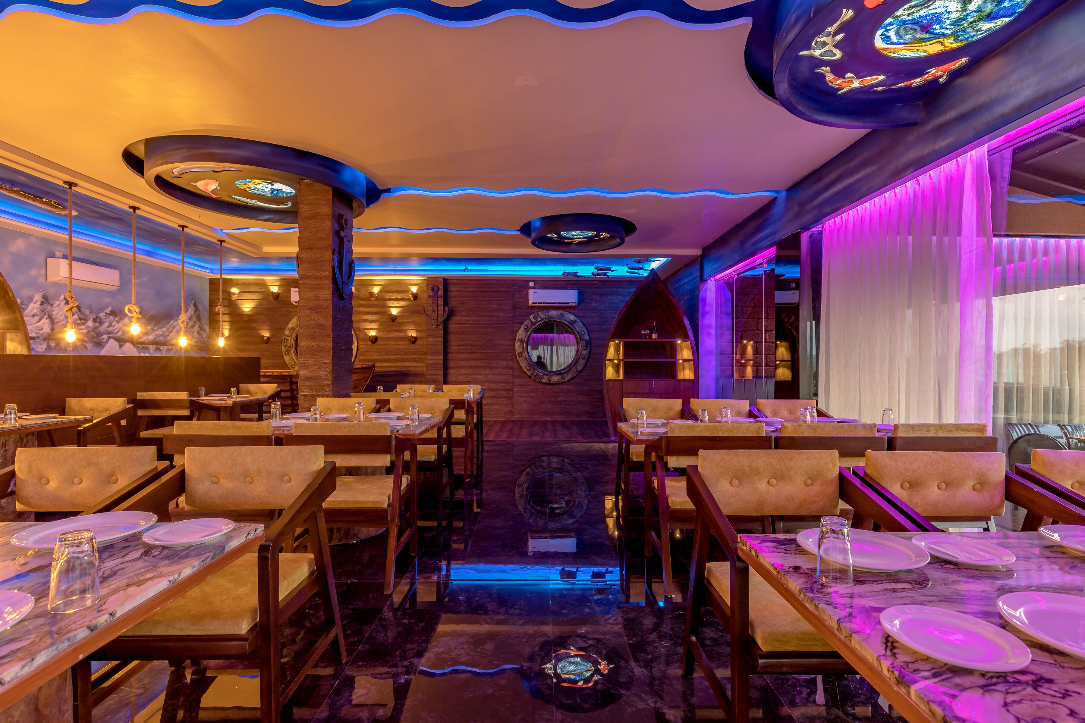 A restaurant with wooden tables and chairs, a central decorative wall, and a ceiling with blue lighting.
