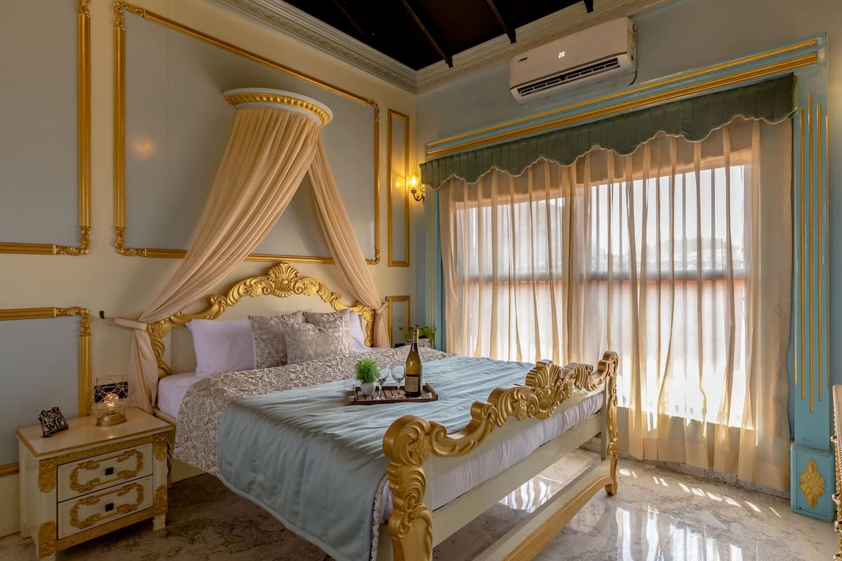 A luxurious bedroom with a four-poster bed, a nightstand with drawers, a bedside table with a lamp, and a large window with curtains.