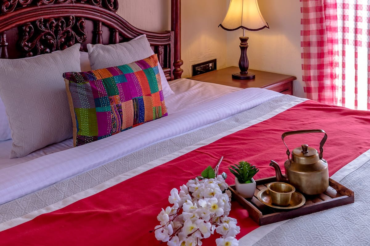 A neatly made bed with a red and white checkered curtain, a wooden headboard, and a bedside table with a lamp and a tray with a teapot and flowers.
