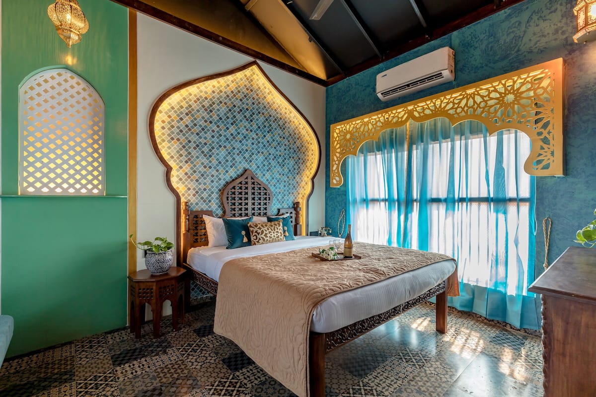 A bedroom with a large bed, a decorative headboard, a small table with a plant, and a window with curtains.