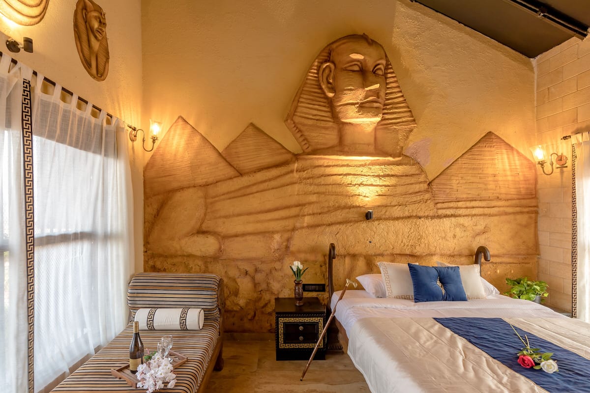 A room with a large wall mural of a pharaoh, a bed with white bedding, a striped sofa, and a small table with a bottle and flowers.