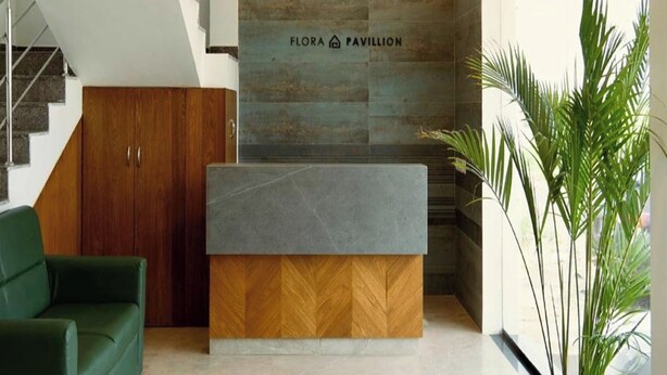 A reception desk with a wooden base and a grey top, a wooden wardrobe, and a wall with the text "FLORA PAVILLION". - Flora Pavillion Near Global Hospital Perumbakkam (Chennai)