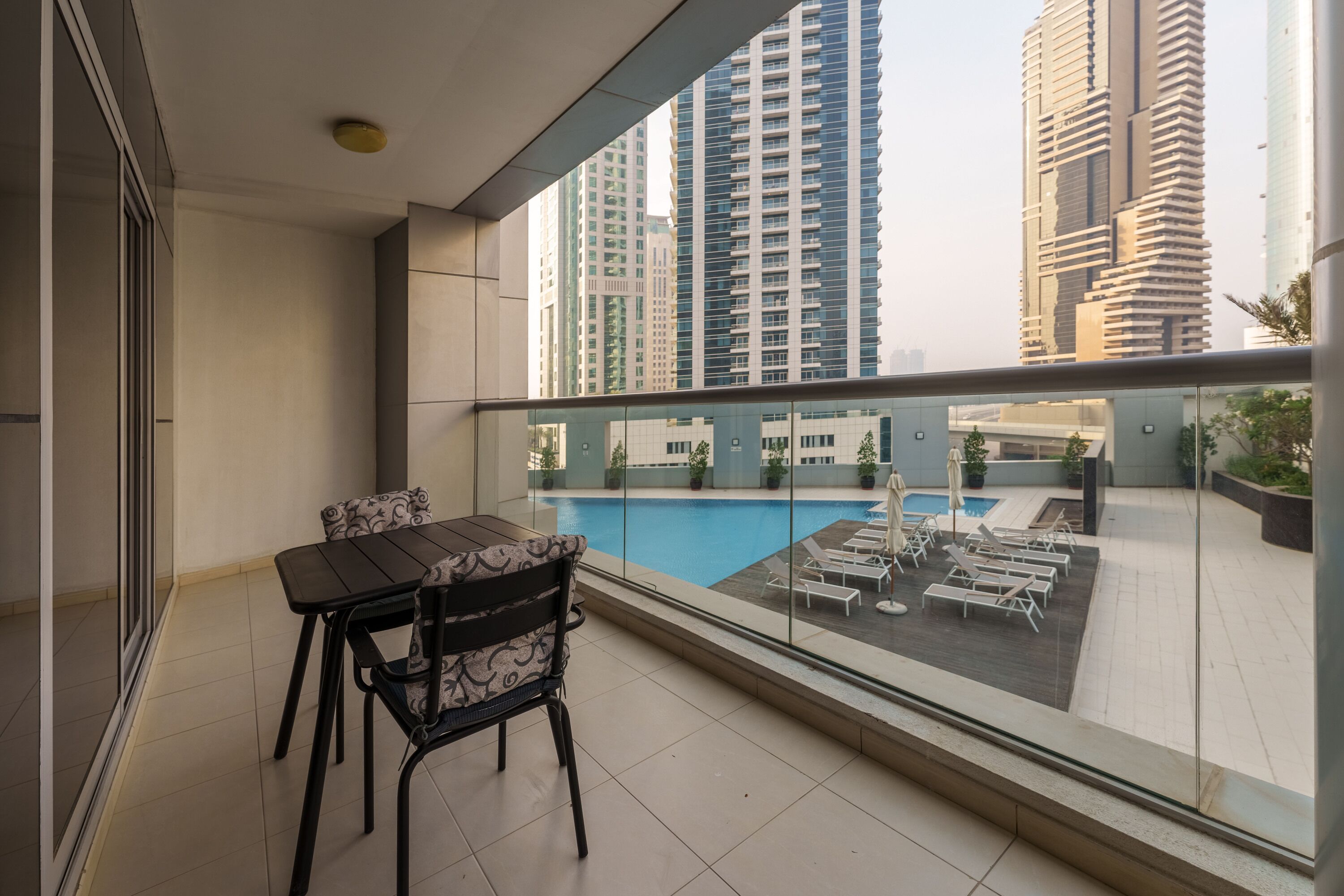 A balcony with a dining table and chairs, overlooking a swimming pool area with lounge chairs and potted plants.