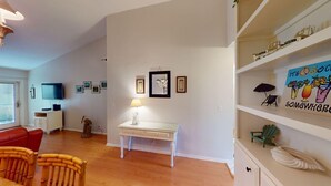 A living room with a white desk, a red sofa, and a television. There are framed pictures on the wall and a shelf with decorative items.