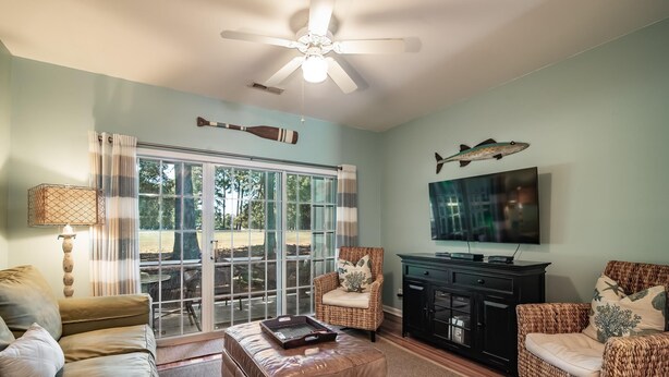 A living room with a ceiling fan, a large window with curtains, a television, and wicker furniture. - River Club 3 Bedroom: Golf Views  and Amenities (Pawleys Island)