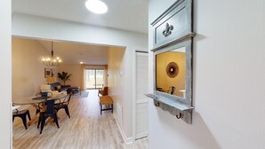 Amplia sala de estar con mesa de comedor, sillas y sofá. Hay un ventanal con cortinas, un ventilador de techo y un espejo decorativo en la pared.