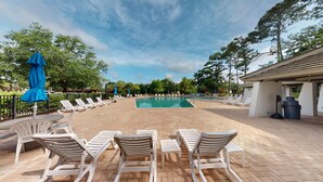 Un área de piscina con sillones de descanso y sombrillas, rodeada de árboles y una zona de estar techada.