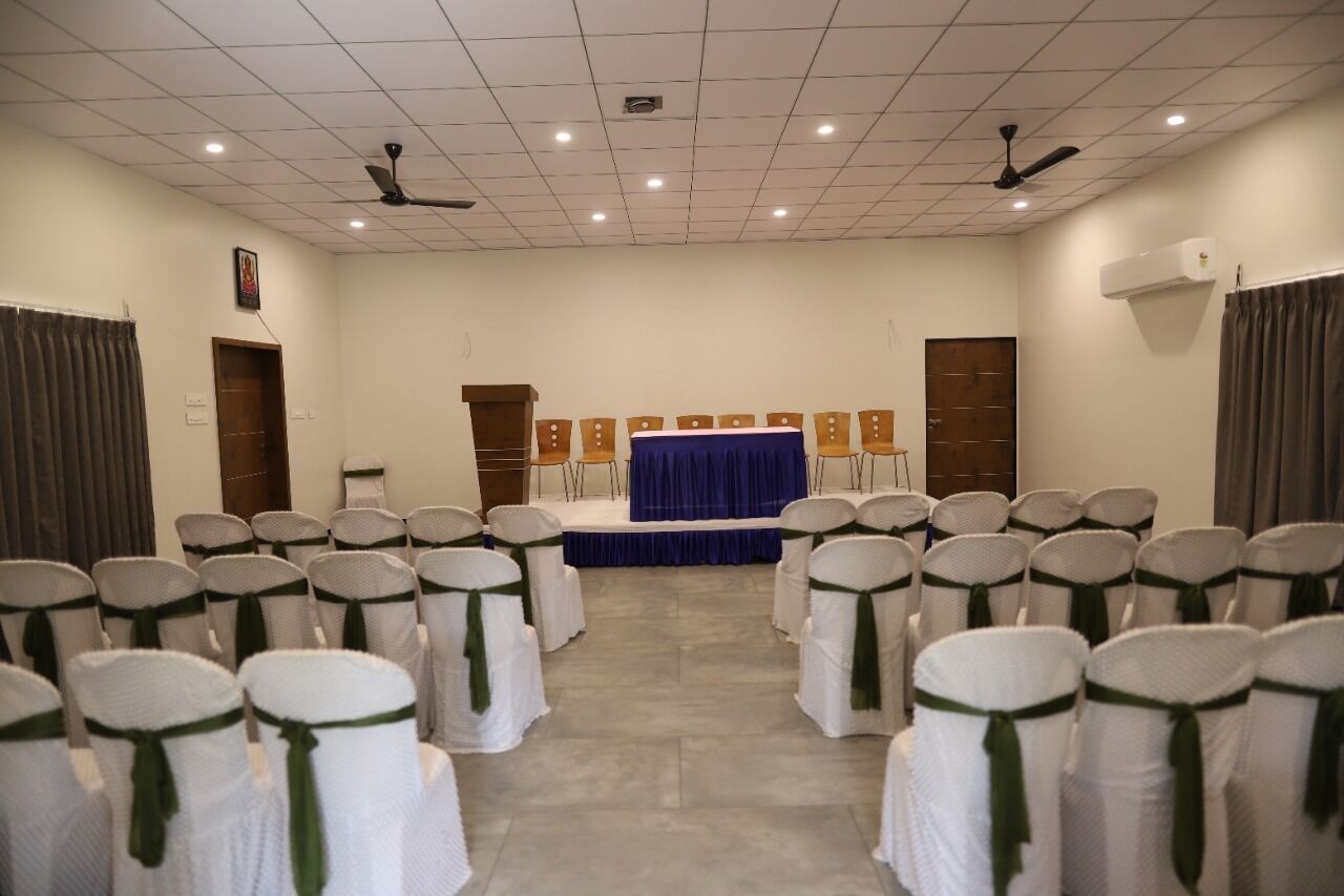Une salle de conférence avec des rangées de chaises blanches ornées de rubans verts, une scène bleue et des ventilateurs de plafond.