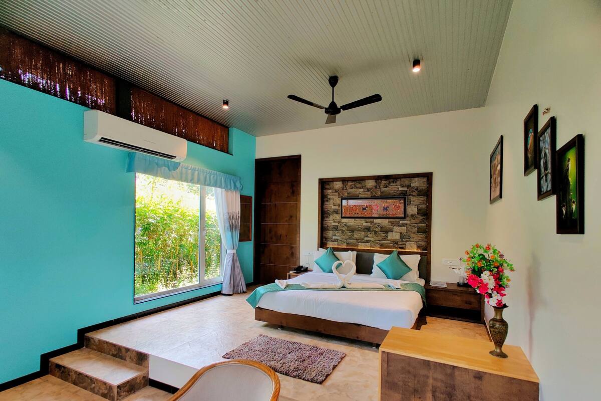 A bedroom with a large window, a bed with pillows, a wooden headboard, a ceiling fan, and a small wooden table with a vase.