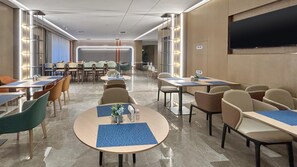 A hotel dining area with round tables, chairs, and a large flat-screen TV mounted on the wall.