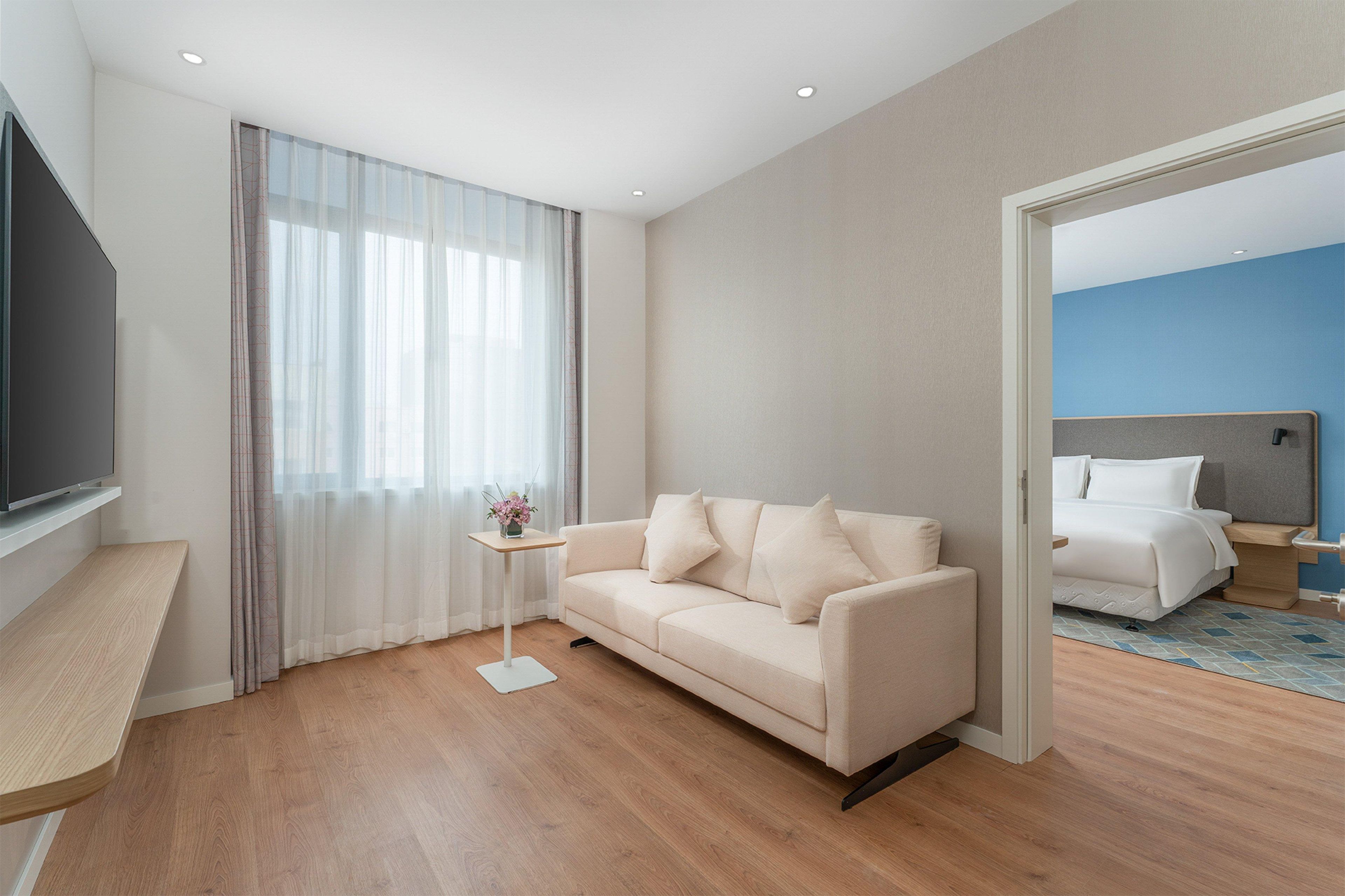Una habitación de hotel moderna con un sofá, un televisor, una pequeña mesa con un florero y vistas a otra habitación con una cama.