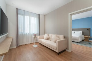 Una habitación de hotel moderna con un sofá, un televisor, una pequeña mesa con un florero y vistas a otra habitación con una cama.