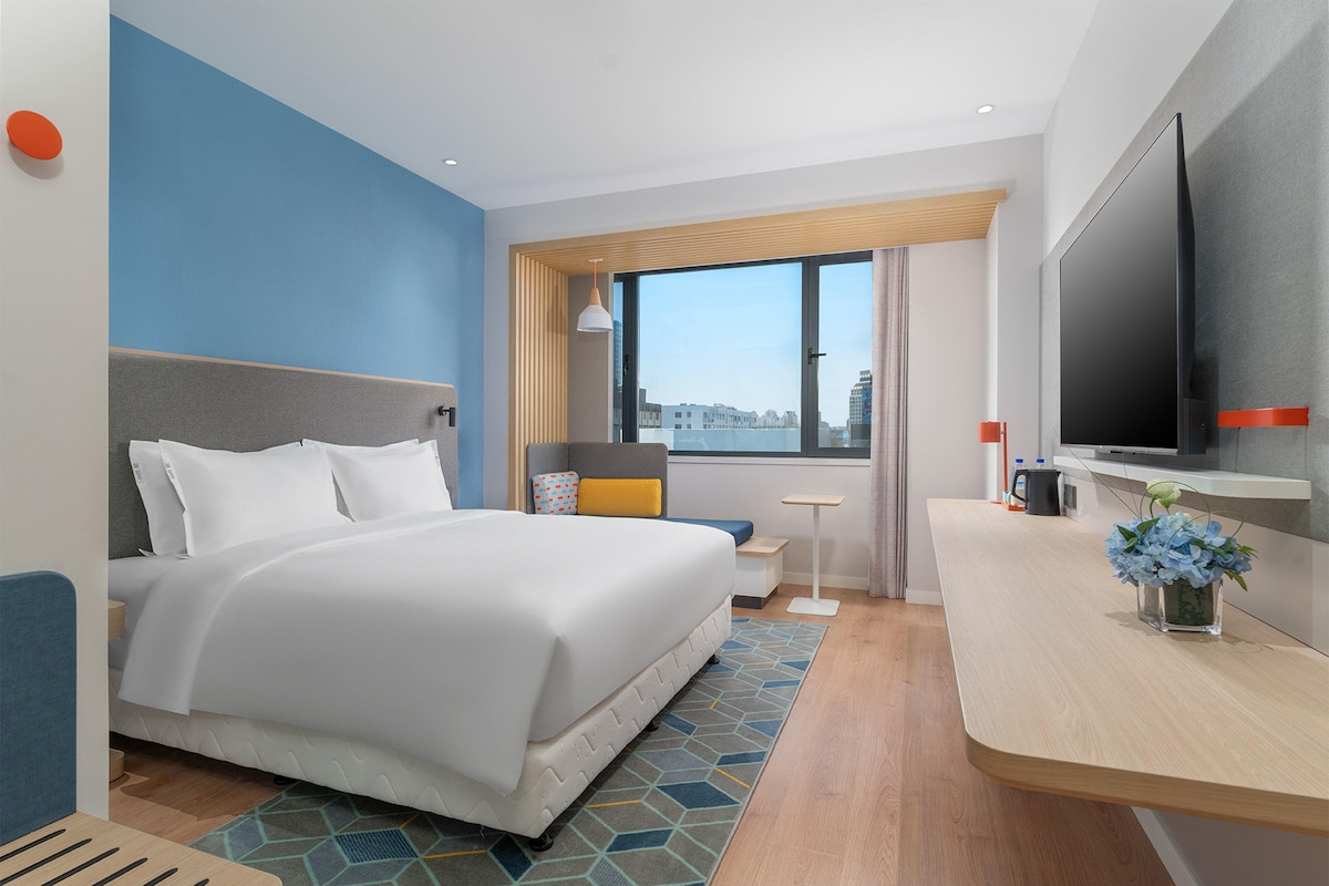 A hotel room with a bed, a desk, a TV, and a window with a city view.