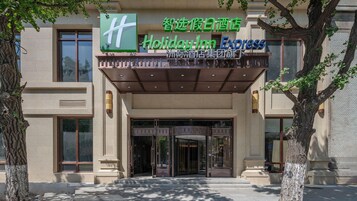 Entrada de hotel con cartel de Holiday Inn Express, un acceso cubierto y un gran árbol a la izquierda.