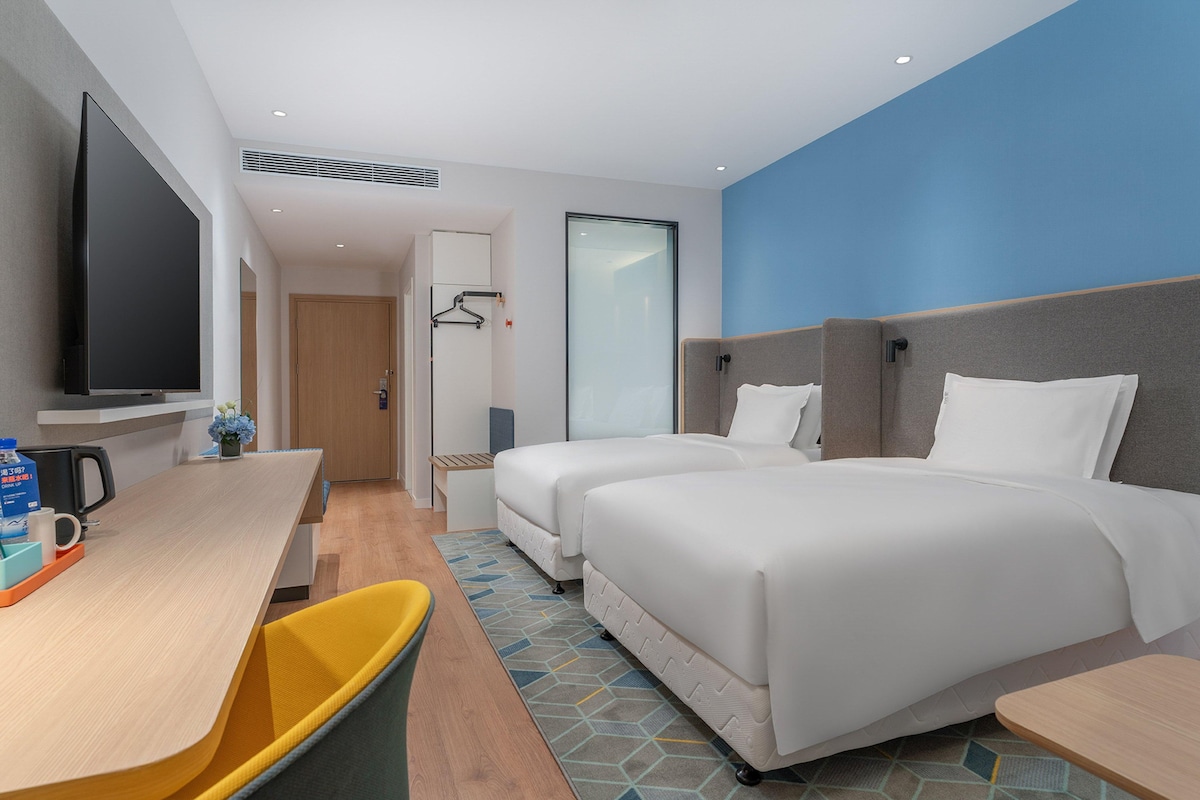 A hotel room with a flat-screen TV, two beds, a desk with a chair, and a trash bin.