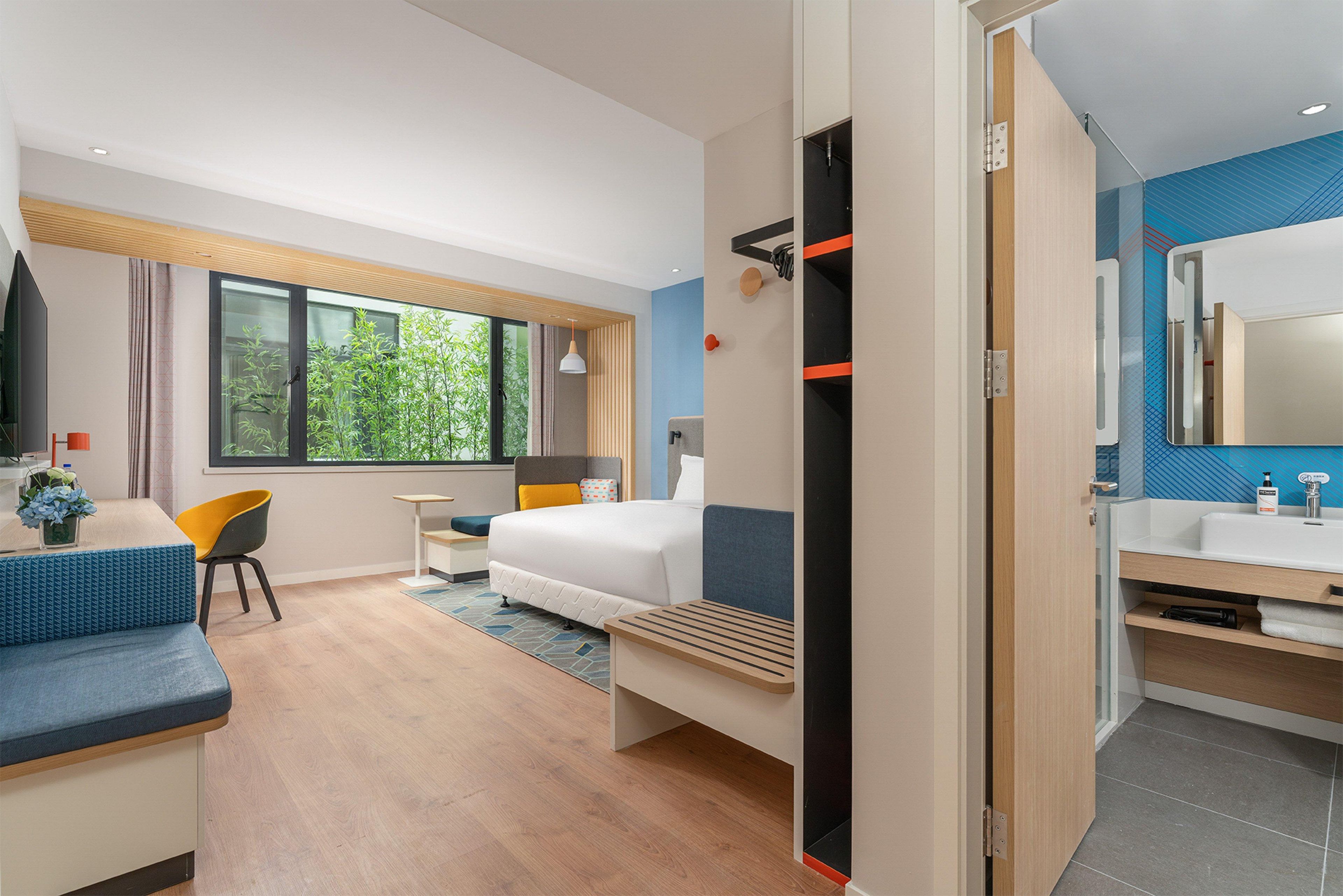 A modern hotel room with a bed, a desk, a chair, a small table, a bathroom with a mirror, and a view of greenery outside.