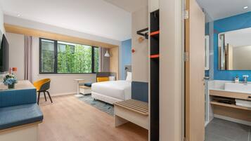 Et moderne hotelværelse med seng, skrivebord, stol, lille bord, badeværelse med spejl og udsigt til grønne områder.