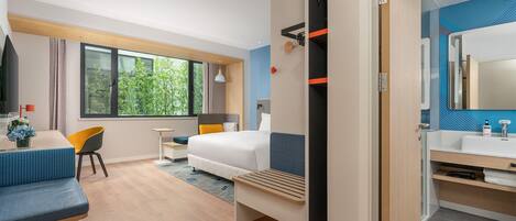 A modern hotel room with a bed, a desk, a chair, a small table, a bathroom with a mirror, and a view of greenery outside.