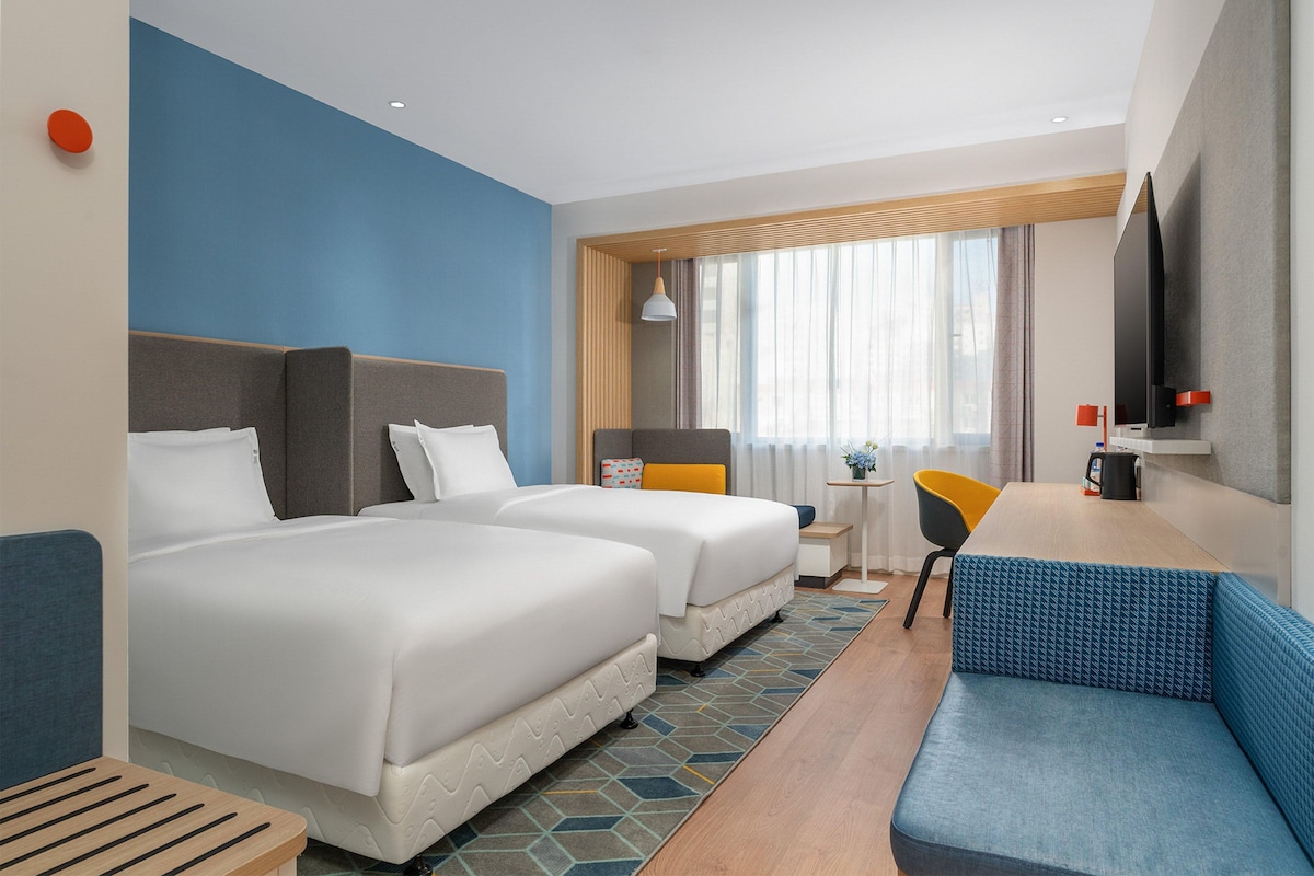 A hotel room with two beds, a sofa, a small table, a chair, a TV, and a window with curtains.