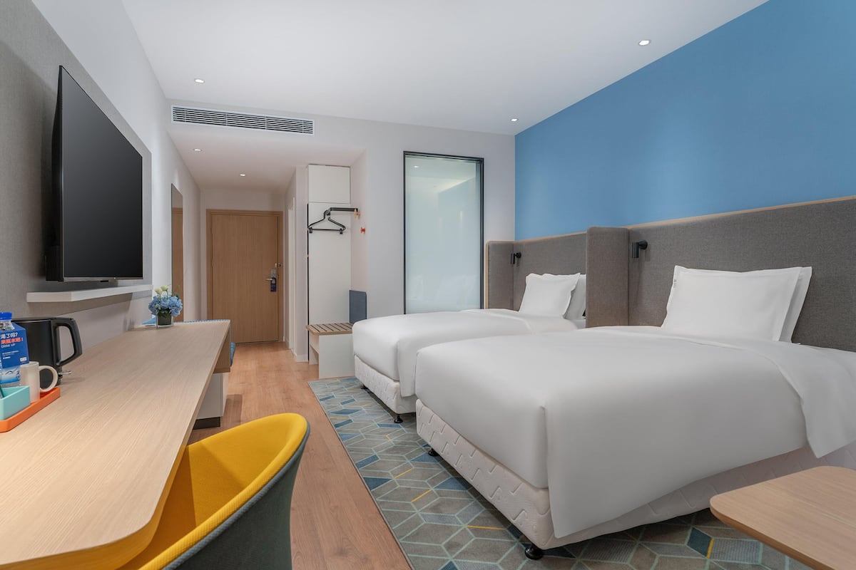 A hotel room with a flat-screen TV, two beds, a desk with a chair, and a trash bin.