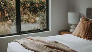 A bedroom with a bed, a window with a view of plants, a bedside lamp, and a pillow. - Tranmere Seaside Serenity (Tranmere)