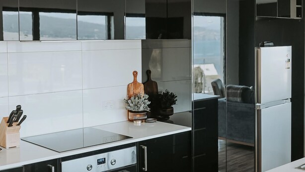 A modern kitchen with a sleek black countertop, stainless steel appliances, and a view of the outdoors through large windows. - Tranmere Seaside Serenity (Tranmere)