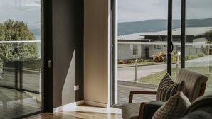 A modern living room with a large glass door leading to a balcony, offering a view of a residential area and mountains. - Tranmere Seaside Serenity (Tranmere)