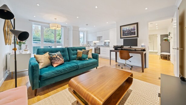 Een ruime woonkamer met een blauwgroene bank, een houten salontafel en een eettafel met stoelen. Achtergrond toont een keuken met witte kasten en een groot raam.